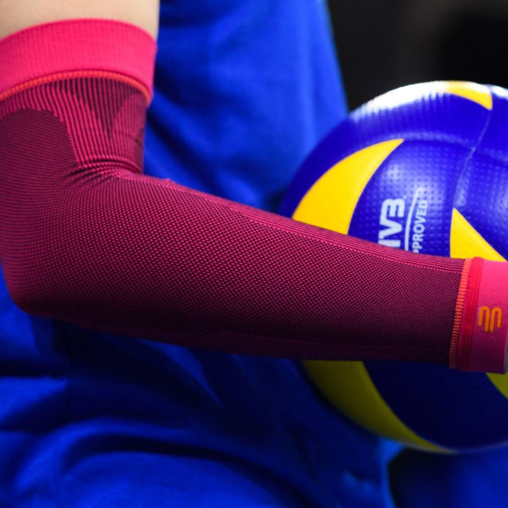 Nahaufnahme eines pinkfarbenen Volleyball-Armsleeves an einem Spielerarm mit einem gelb-blauen Volleyball im Hintergrund.