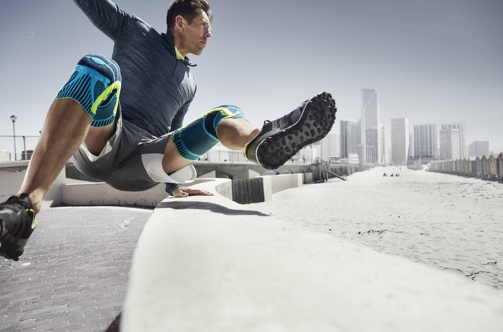 Mann mit zwei Kniebandagen springt beim Urban Running über ein Hindernis	 im Hintergrund Wolkenkratzer