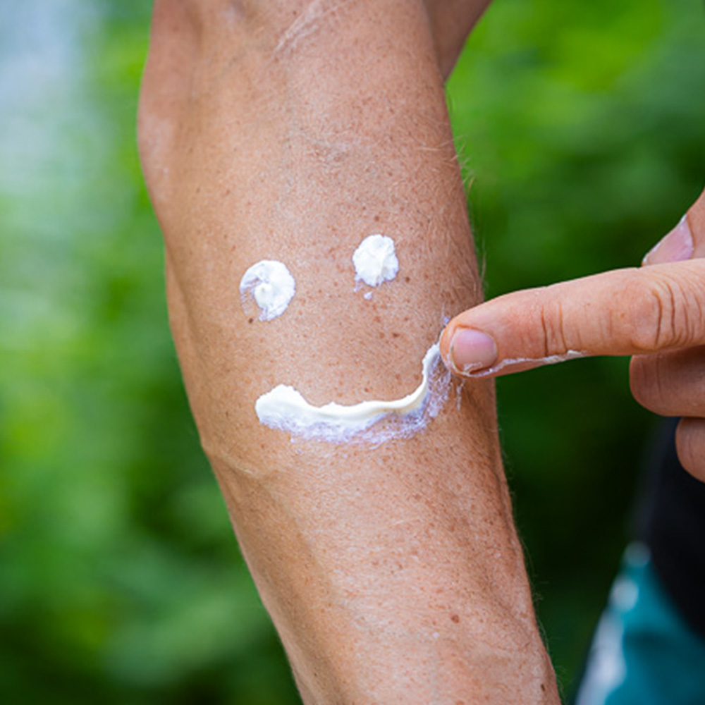 Bauerfeind Sports Sonnenschutzcreme einfach auf den Arm aufgetragen und ein lächelndes Gesicht mit Creme formen Auf einem Unterarm ist Sonnenschutzcreme von Bauerfeind Sports aufgetragen, die ein lächelndes Gesicht bildet. Eine Hand zeigt auf den "Mund" des Smileys. Die Szene findet im Freien statt, umgeben von grüner Natur.