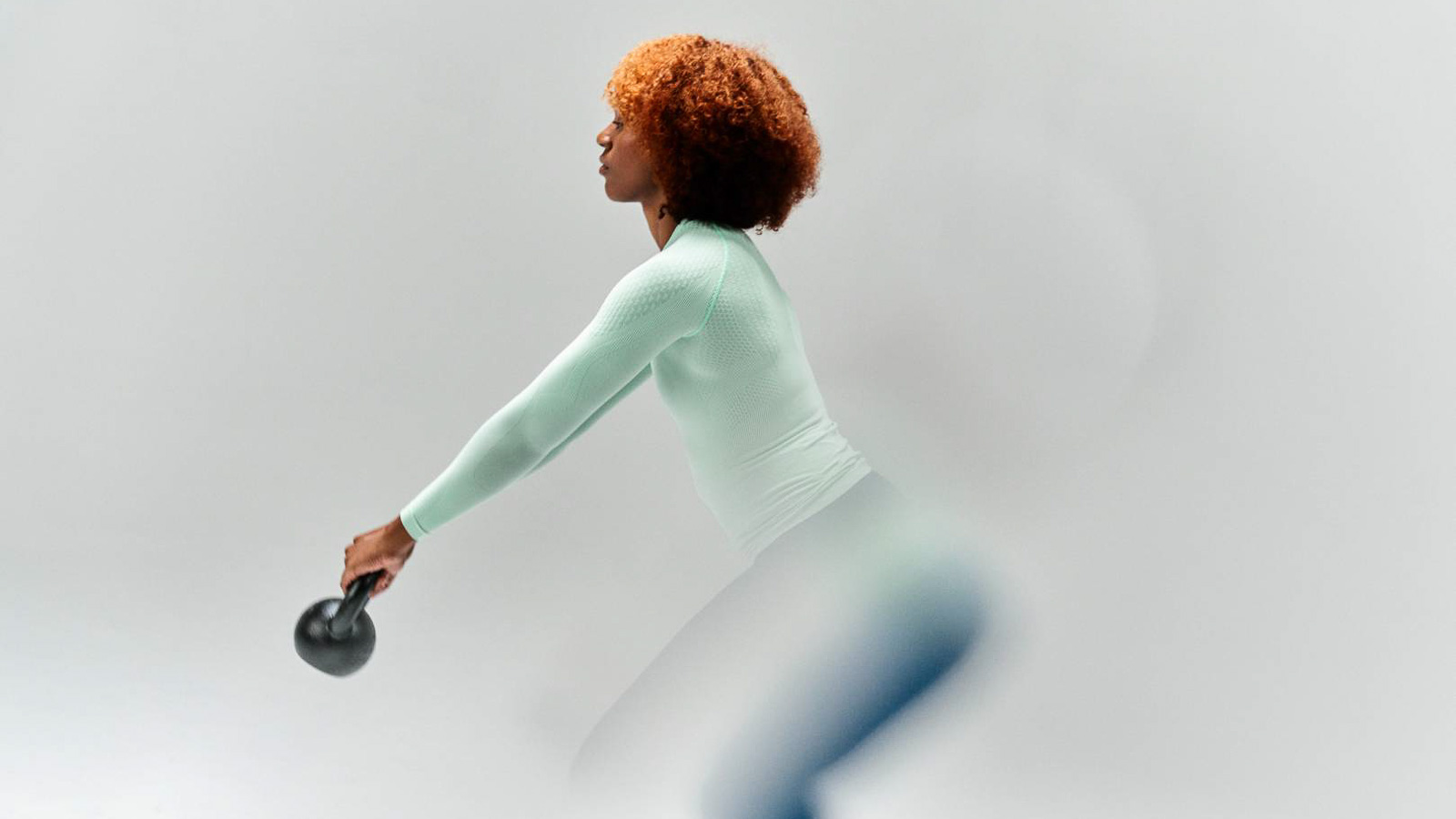 Effektives Training mit Stil - Kettlebell-Übungen in hochwertiger Kompressions-Sports-Wear! Eine Frau in einem mintfarbenen Kompressions-Sports-Wear-Shirt führt eine Kettlebell-Übung durch. Der Fokus liegt auf ihrer Bewegung und der funktionalen Sportswear vor einem neutralen Hintergrund.