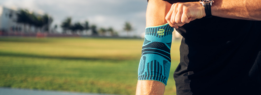Bereit für deinen großen Wurf! Nahaufnahme eines Sportlers, der sich konzentriert eine blau-schwarze Ellenbogenbandage am rechten Arm anlegt. Im Hintergrund ist eine sonnige Sportanlage zu sehen.