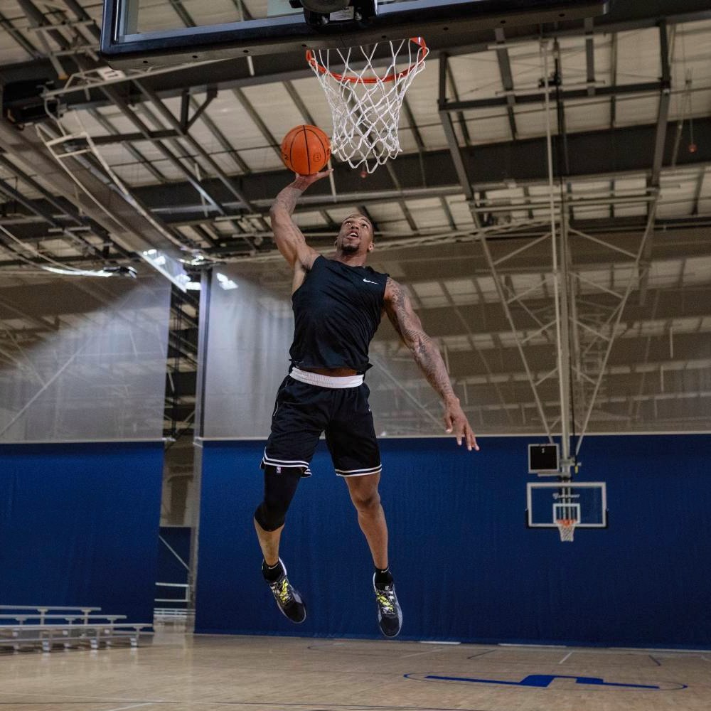 Hoch hinaus – mit voller Power zum Dunk! Ein Basketballspieler in einem schwarzen Outfit springt hoch in Richtung Korb für einen Dunk. Er trägt eine schwarze Kniebandage an seinem rechten Bein, die während des Spiels für Stabilität sorgt. Die Sporthalle im Hintergrund ist hell beleuchtet.