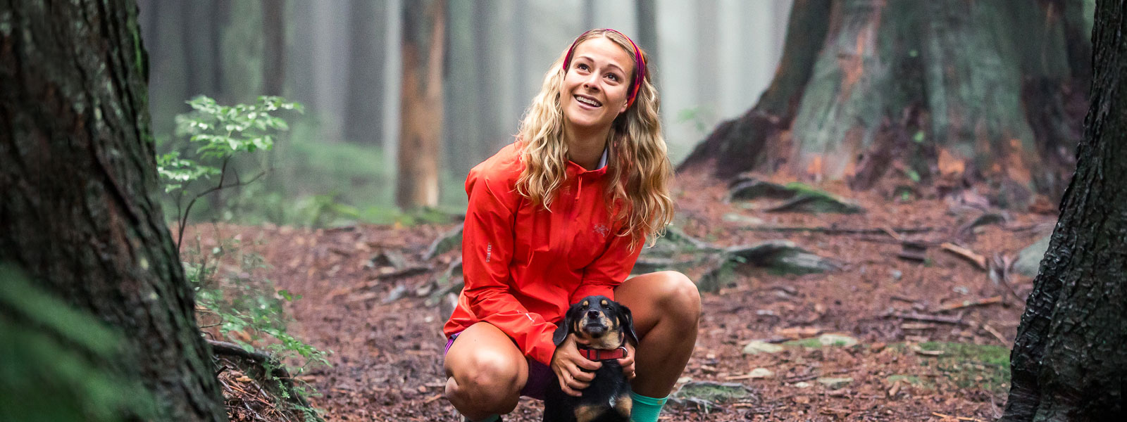 Waldidyll mit Hund: Hailey van Dyk bei einer kleinen Laufpause Frau mit roter Laufjacke und Stirnband hält ihren kleinen Hund auf einem Waldweg und lächelt