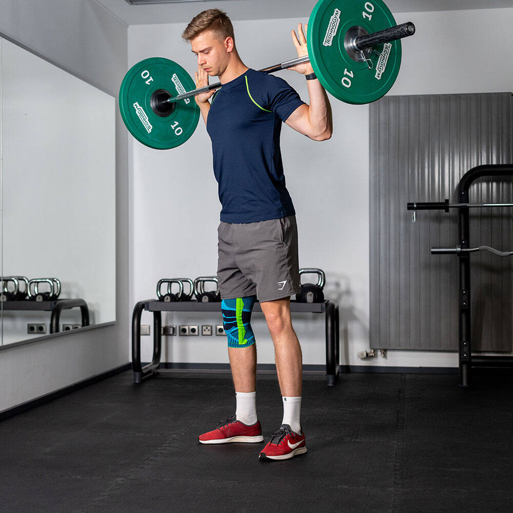 Mann mit Kniebandage in Fitnessstudio hält eine Langhantel in seinem Genick vor der Ausführung eines Squats