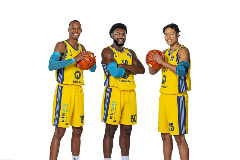 Drei Basketballspieler von Alba Berlin in ihren gelben Team-Trikots posieren mit Basketbällen vor einem neutralen weißen Hintergrund. Jeder Spieler lächelt und hält einen Basketball mit entspanntem, professionellem Ausdruck.