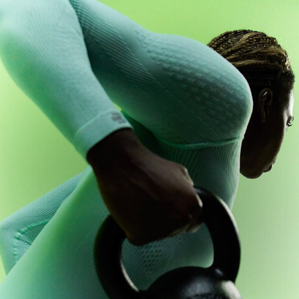 Athletin in mintgrünem Kompressionsoberteil und dunkelblauer Kompressionshose macht Armtraining Athletin in mintgrünem Kompressionsoberteil und dunkelblauer Kompressionshose streckt ihren Arm nach oben, während sie eine Hantel in der Hand hat