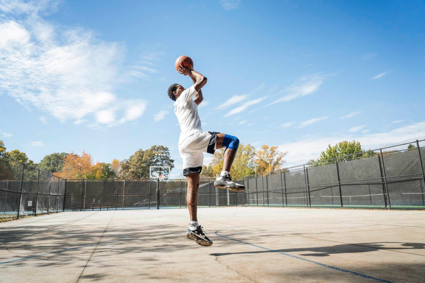 Basketballer springt auf einem Streetcourt für den Wurf ab	 Hände mit Ball überm Kopf	 ein Bein angewinkelt	 das andere senkrecht nach unten zeigend