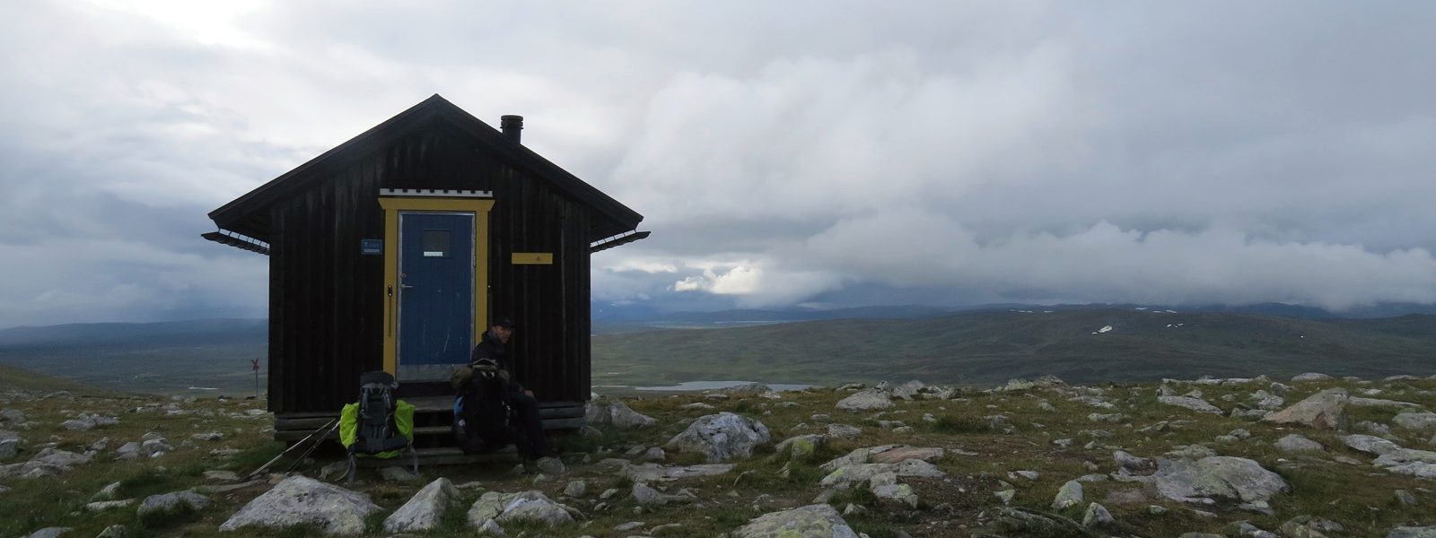 Eine einsame Hütte im Gebirge inmitten von begrasten Wiesen mit kleinen Felsen	 im Hintergrund die dichte Wolkendecke und ein kleiner Bergsee