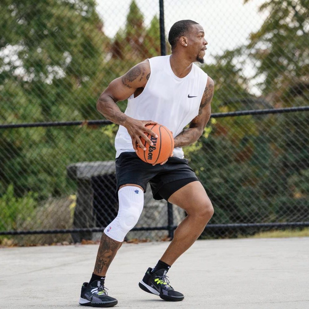 Stabilität und Style in jedem Spiel Basketballspieler im weißen Trikot bereitet sich auf einen Wurf vor. Sein rechtes Knie ist mit einer weißen Kompressionsbandage stabilisiert, er steht auf einem Freiplatz mit Körben im Hintergrund.