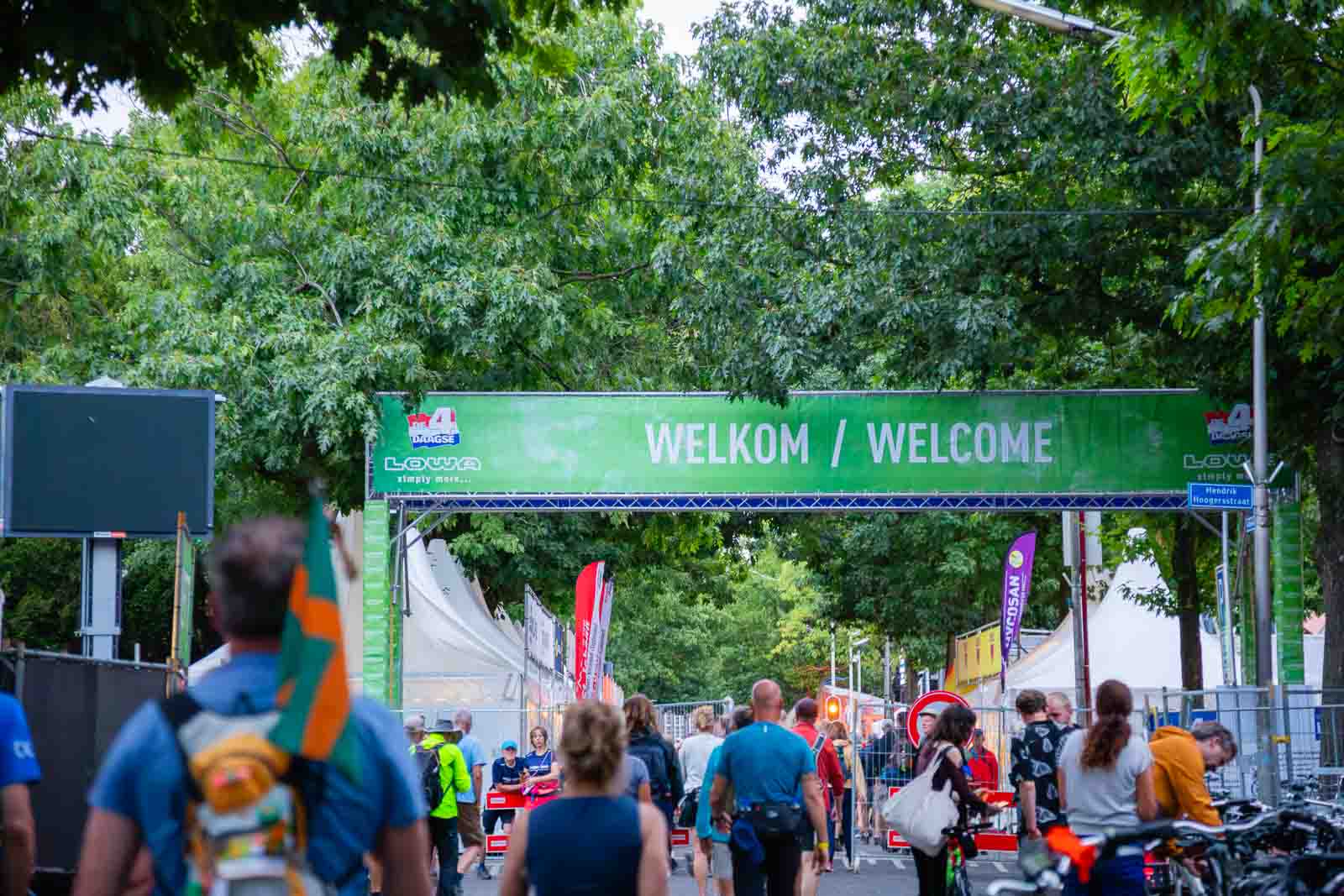 Eingang zum 4Daagse Event in Nijmegen mit grünem Banner „Welkom / Welcome“ und zahlreichen Wanderern, die zum Startbereich gehen.