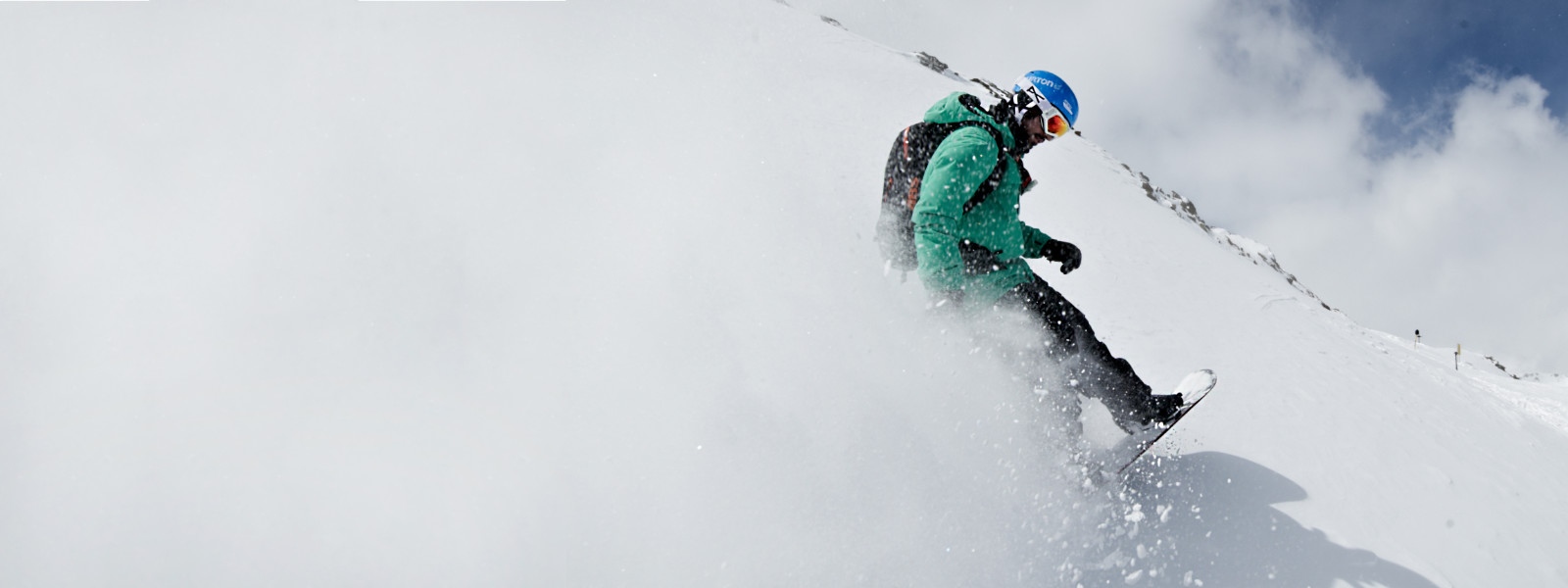 Snowboarder mit grüner Jacke wirbelt beim Bremsen Schnee auf	 der die komplette linke Bildseite weiß färbt