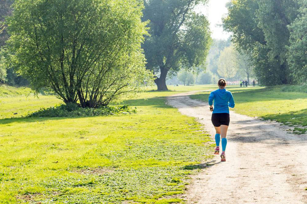 Frau mit blauem Pullover und blauen Waden-Sleeves läuft durch den Park	 Rückansicht