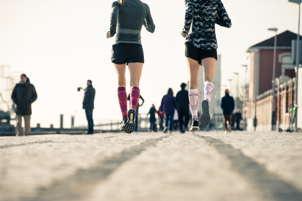 Froschperspektive: Läuferinnen mit lilafarbenen und weißen Laufsocken laufen durch eine belebte Stadt	 im Hintergrund Menschen	 Häuser und Laternen