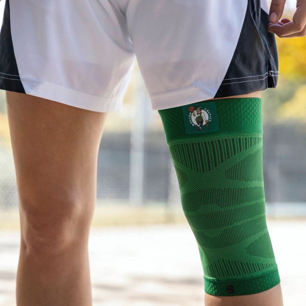 Boston Celtics Support – Stabilität für das Spiel Nahaufnahme eines Spielers mit einer grünen Boston Celtics-Kniebandage auf einem Basketballplatz. Die Bandage zeigt das Teamlogo und passt sich ergonomisch an das Knie an.