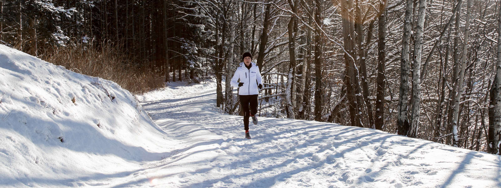 Mit der richtigen Ausrüstung ist das Laufen im Winter (mindestens) genauso schön wie in den anderen Jahreszeiten Frau in weißer Laufjacke joggt auf einem schneebedeckten Weg zwischen Bäumen und sieht in Richtung Kamera