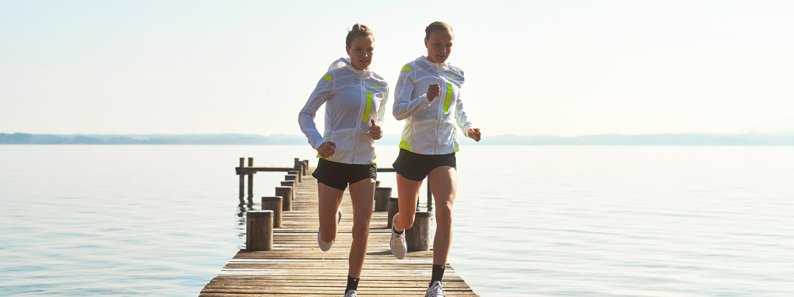 Zwei Läuferinnen in weißen Laufjacken und mit kurzen Hosen laufen über einen Steg in Richtung Ufer