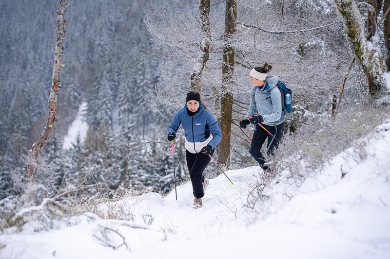 Mit Stöcken durch den Schnee – Winterwandern für Gipfelstürmer! Zwei Frauen wandern mit Trekkingstöcken durch eine verschneite Winterlandschaft. Sie tragen warme Funktionskleidung, eine blaue Jacke und eine hellblaue Jacke, sowie Handschuhe und Stirnbänder.