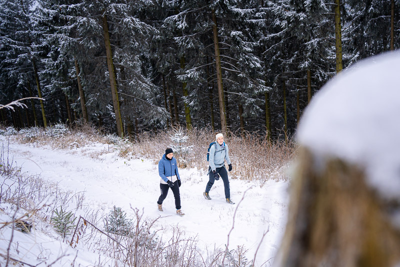 Winterwandern – Wo jeder Schritt ein knirschendes Abenteuer ist. Zwei Frauen wandern durch eine verschneite Waldlandschaft. Sie tragen warme Winterkleidung und nutzen Wanderstöcke, während sie einen Pfad entlanglaufen. Im Hintergrund sind schneebedeckte Bäume und ein dichter Wald zu sehen.