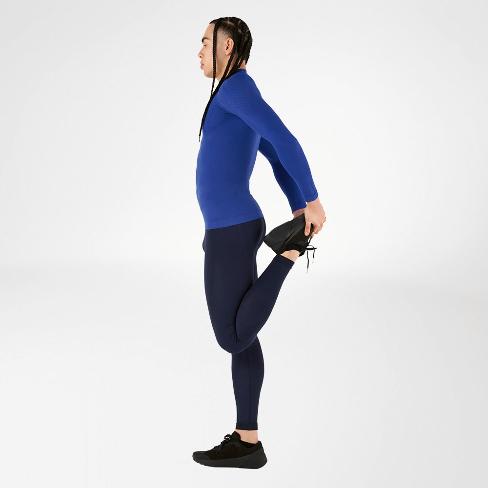 Ein Sportler in einem blauen Kompressionsoutfit macht eine Dehnübung und hält dabei ein Bein nach hinten, vor einem weißen Hintergrund. Das Outfit bietet hohen Tragekomfort und optimale Unterstützung der Muskulatur.