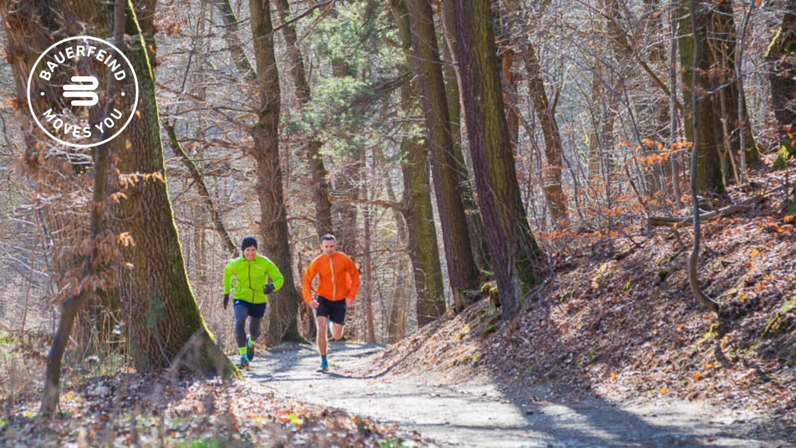 Geschwindigkeit pur – Mit Tempo durch den Wald Zwei Männer in leuchtenden Sportjacken laufen mit hohem Tempo auf einem Waldweg entlang. Die dichten Bäume und das Sonnenlicht sorgen für eine lebendige Atmosphäre, während beide Sportler dynamisch unterwegs sind.