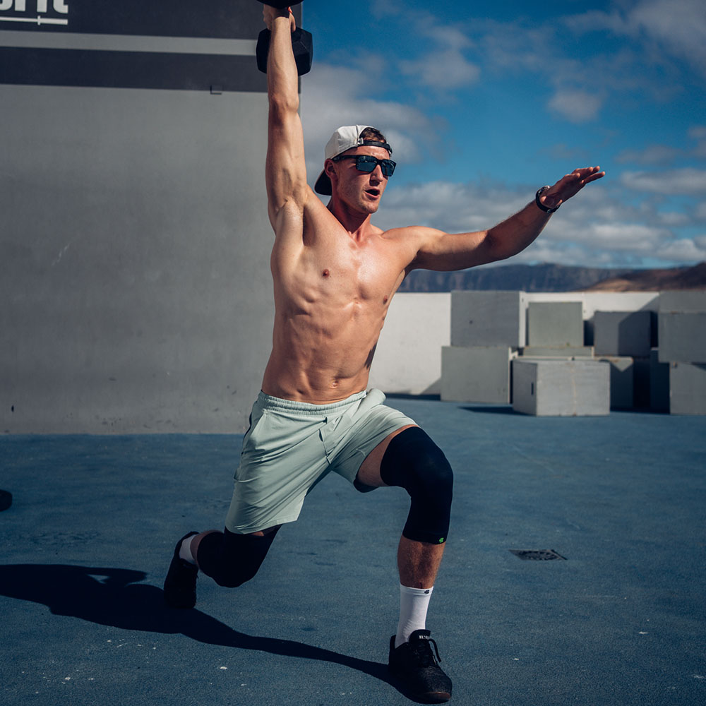 Maximale Stabilität – auch bei intensivem Training! Ein sportlicher Mann führt eine einbeinige Kniebeuge mit einer Hantel über dem Kopf aus, während er eine schwarze Kniebandage trägt.