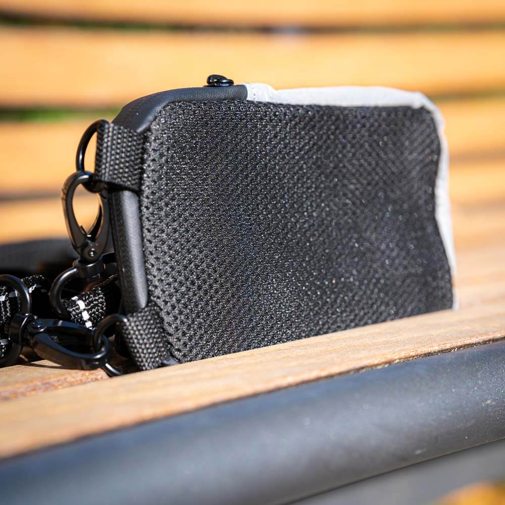 Robustes Design: Rückseite der Bauerfeind Sports Tasche mit Netzmaterial Nahaufnahme der Rückseite der grauen Bauerfeind Sports Tasche, die mit einem atmungsaktiven schwarzen Netzmaterial ausgestattet ist. Die Tasche liegt auf einer Holzbank und hat zwei Karabiner zur Befestigung.