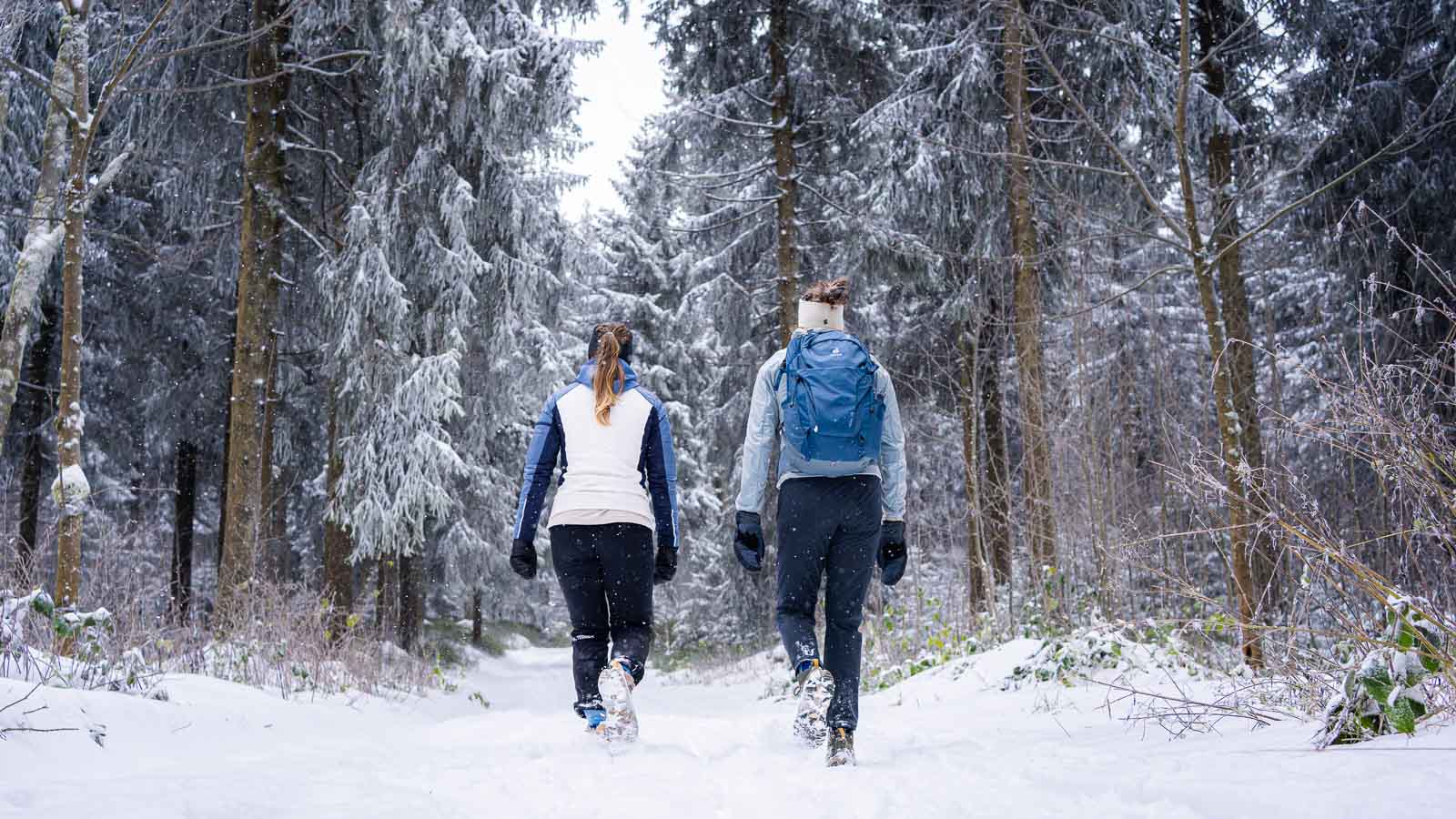 Winterwandern – Wo jeder Schritt ein knirschendes Abenteuer ist Zwei Frauen wandern durch eine verschneite Waldlandschaft. Sie tragen warme Winterkleidung und nutzen Wanderstöcke, während sie einen Pfad entlanglaufen. Im Hintergrund sind schneebedeckte Bäume und ein dichter Wald zu sehen.