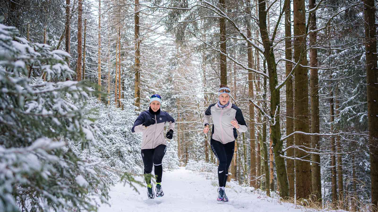 Zwei Personen joggen auf einem schneebedeckten Waldweg. Die verschneiten Tannen bilden einen dichten, winterlichen Hintergrund. Beide Läufer tragen warme Jacken und Stirnbänder für den Winterlauf.