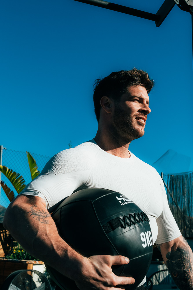 Athlet steht draußen mit einem schwarzen 9-kg-Medizinball im Arm, trägt ein eng anliegendes Sportshirt und blickt im Sonnenlicht nach vorn – funktionelles Krafttraining im Hybrid-Athlet-Kontext.