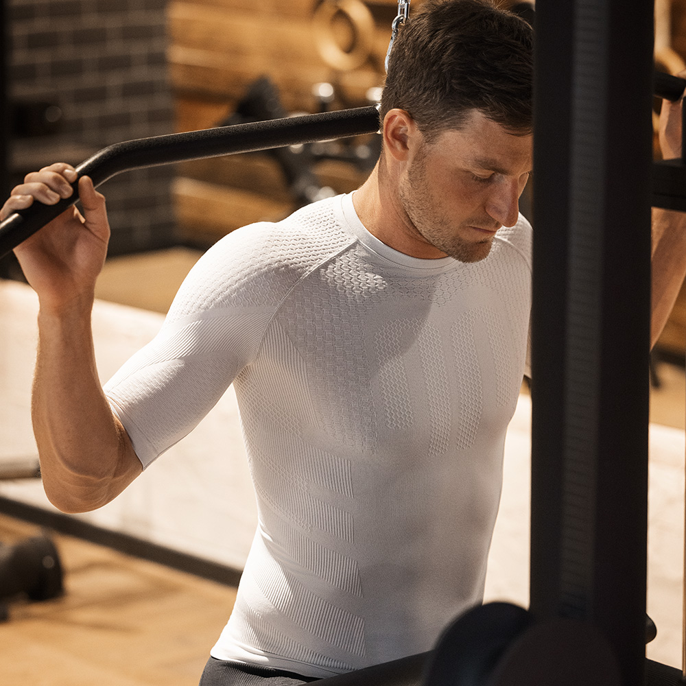 Spüre die Power – Kompression, die dein Workout auf das nächste Level bringt Sportler trägt ein beiges, kurzärmliges Kompressionsshirt während des Trainings an einer Kraftmaschine im Fitnessstudio. Das nahtlose, eng anliegende Material unterstützt die Muskulatur und sorgt für eine stabile Körperhaltung.