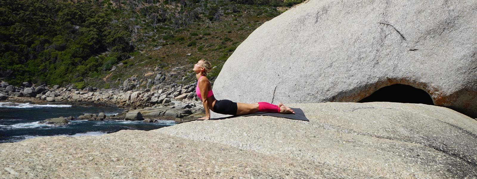 Frau macht Yoga Übungen auf großem Felsen am Wasser und pinke Unterschenkel-Sleeves