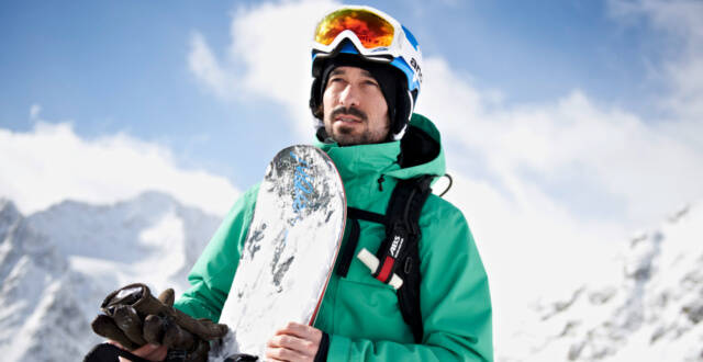 Snowboarder mit grüner Jacke mit Board in der Hand vor Bergkulisse