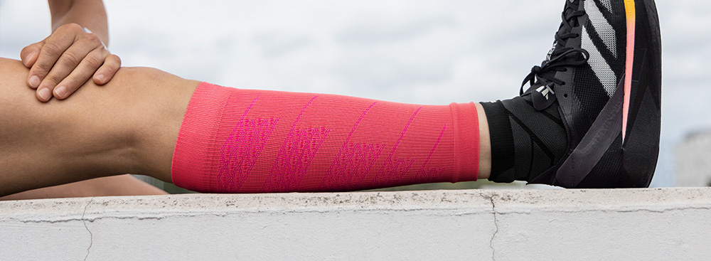 Power in Pink – bereit für den nächsten Run! Ein Sportler sitzt am Boden und dehnt sein ausgestrecktes Bein. Er trägt leuchtend pinkfarbene Compression Sleeves an der Wade, kombiniert mit schwarzen Sportschuhen.