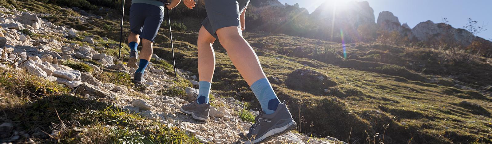 Sicherer Tritt im unwegsamen Gelände: Unsere Outdoor Performance Mid Cut Socks Nahaufnahme Unterschenkel einer Wanderin in Wanderschuhen und mit grünen mittellangen Wandersocken die einen steinigen Weg nach oben geht im Hintergrund ihr Wanderpartner