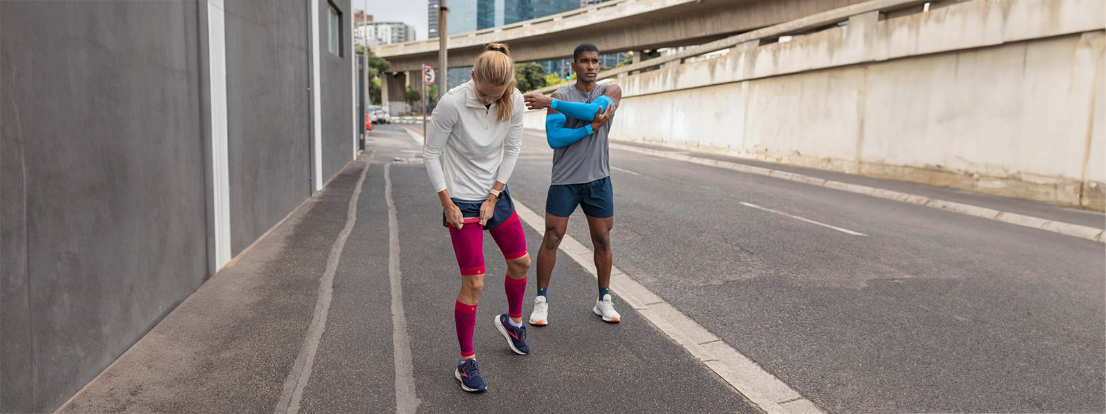 Eine Frau mit pinken Compression Sleeves und ein Mann mit blauen Arm Sleeves und kurzen Laufsocken machen eine Laufpause am Straßenrand einer Großstadt und stretchen sich