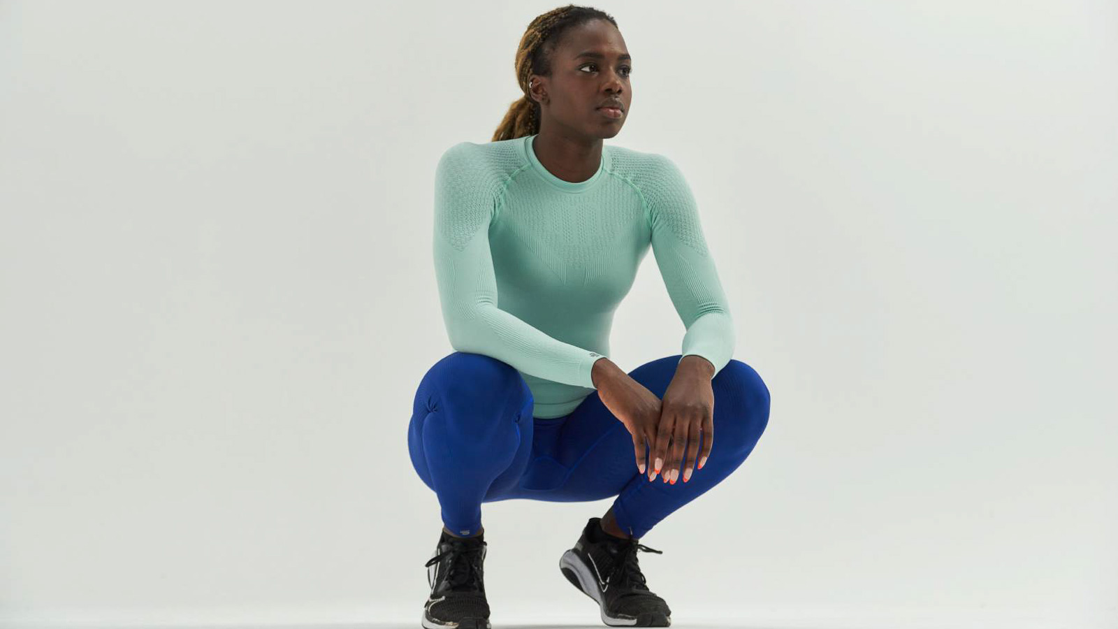 Athletin in mintgrünem Kompressions-Shirt und blauer Kompressionshose Athletin in mintgrünem Kompressions-Shirt und blauer Kompressionshose in Hockstellung vor weißem Hintergrund.