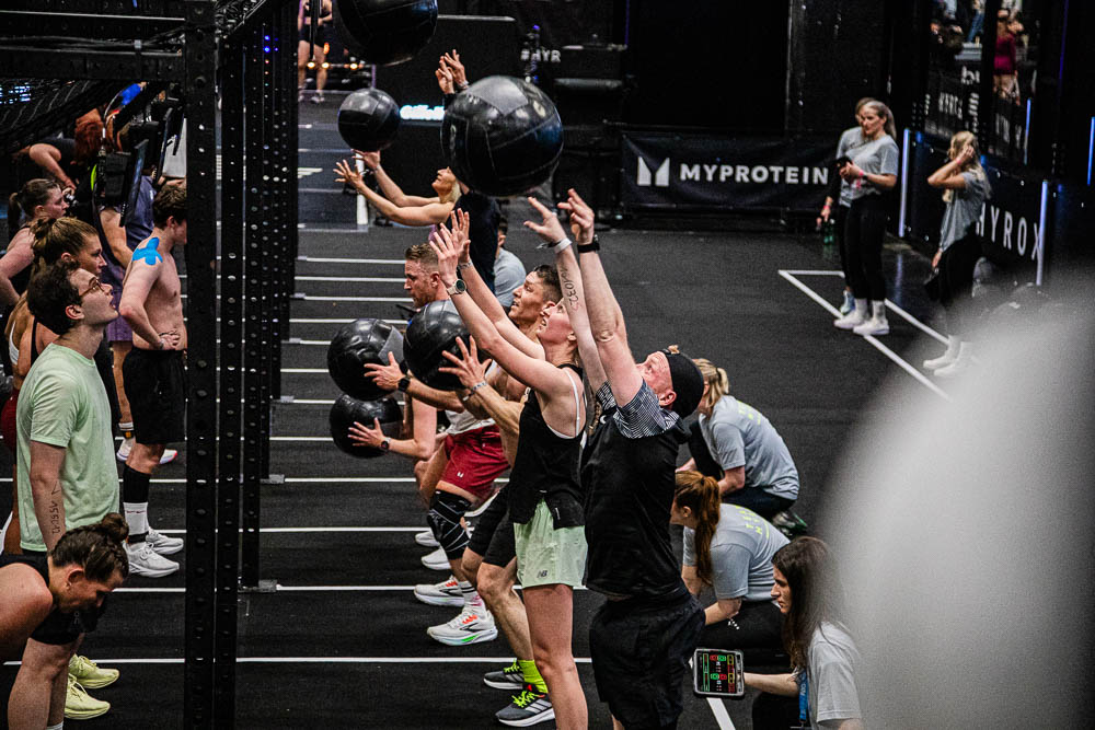 Athletinnen und Athleten absolvieren Wall Balls in einer HYROX-Trainingszone und werfen schwarze Medizinbälle nach oben, während Kampfrichterinnen und Betreuende die Wiederholungen beobachten; die Szene zeigt intensive Ganzkörperarbeit, Konzentration