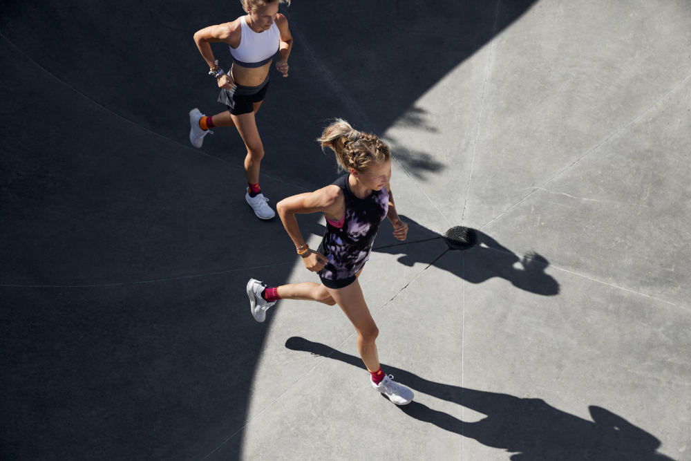 Läuferinnen mit Sprunggelenkbandagen laufen in sommerlichem Outfit über eine Betonfläche	 Perspektive von schräg oben