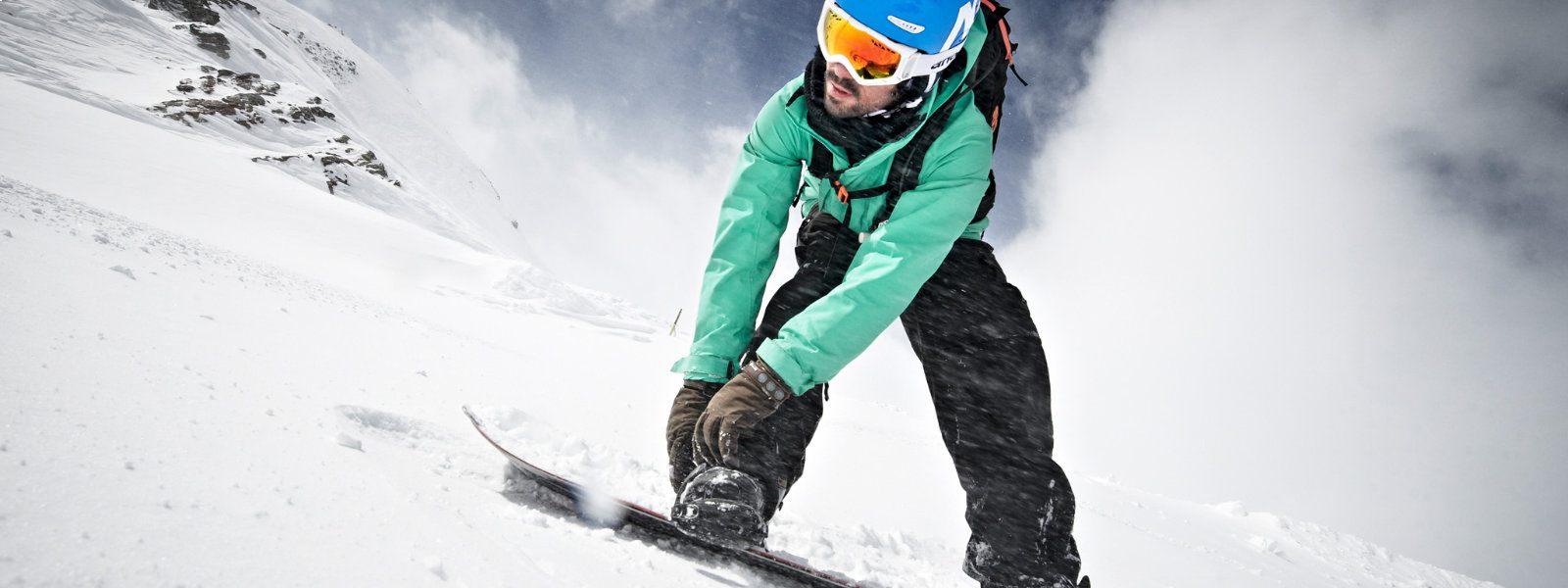 Leichte Froschperspektive: Snowboarder in grüner Jacke befestigt mit seinen Händen eine Bindung und blickt nach unten	 während um ihn herum viel Schnee aufgewirbelt wird 
