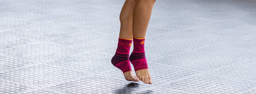 Zoom auf die Beine einer Frau die Sportbandagen für das Sprunggelenk trägt	 sie steht auf den Zehenspitzen auf einem grauen Untergrund