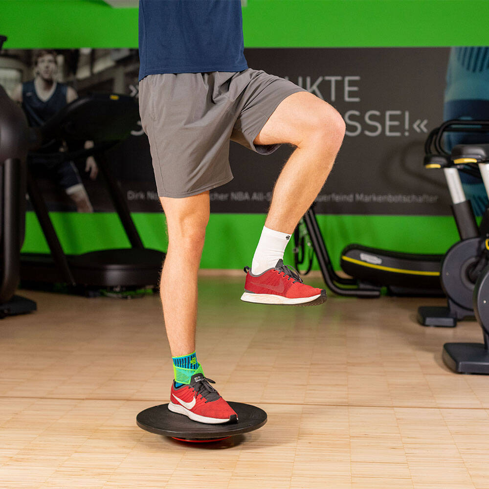 Mann in einem Fitnessstudio balanciert mit dem rechten Bein auf einem Balance Pad und trägt eine Sprunggelenkbandage