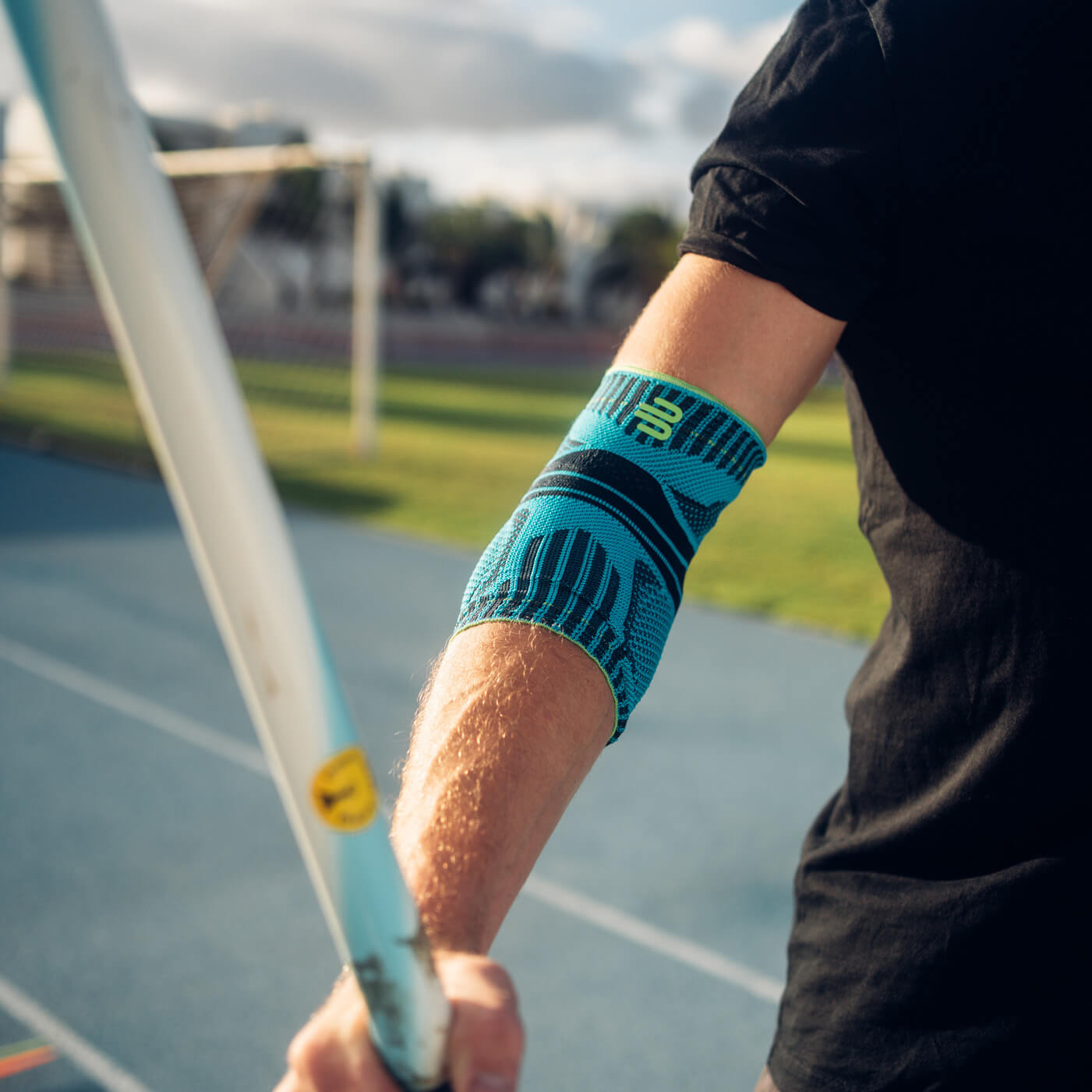 Nahaufnahme eines Arms mit blauer Ellenbogenbandage beim Speerwurf. Im Hintergrund ist eine Laufbahn und eine sonnige Landschaft zu sehen. Präzise und kraftvoll – mit stabiler Unterstützung für deinen Ellenbogen.