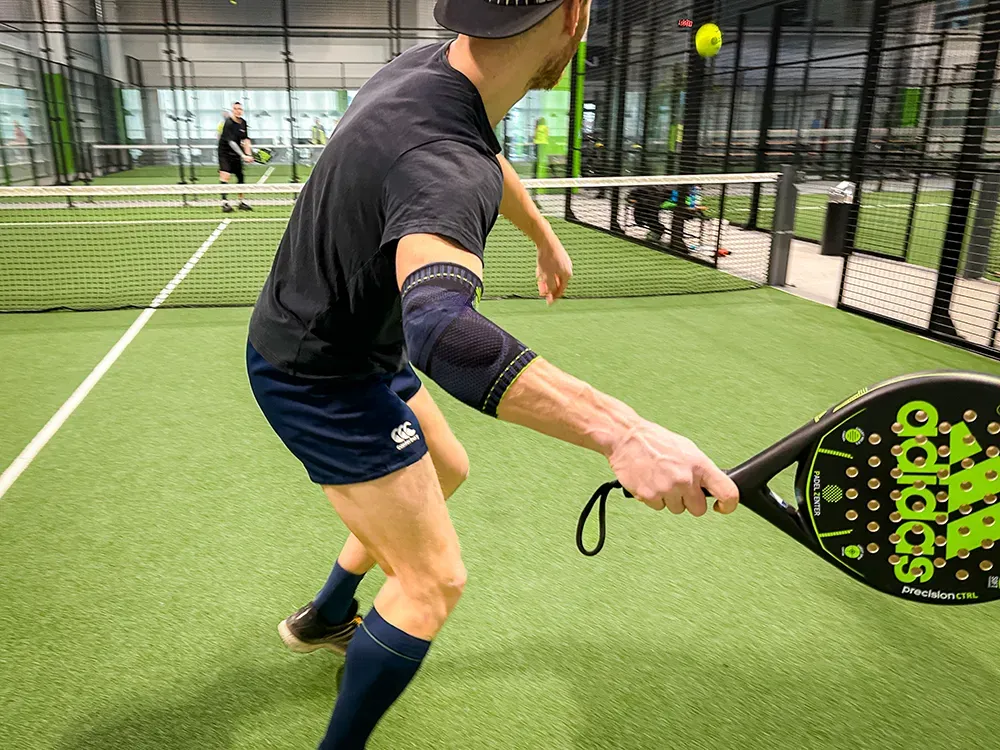 Volle Konzentration beim Padel-Match! Ein Padel-Tennisspieler in schwarzem Shirt und Shorts schlägt den Ball mit voller Konzentration. Am rechten Arm trägt er eine dunkelblaue Sportbandage für zusätzlichen Halt.