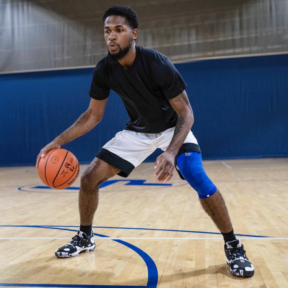 Stabilität auf dem Court – volle Kontrolle beim Spiel. Ein Basketballspieler auf einem Hallenplatz in der Verteidigungshaltung, trägt eine blaue Kompressions-Kniebandage am rechten Bein. Der Holzboden reflektiert das Licht der Halle.