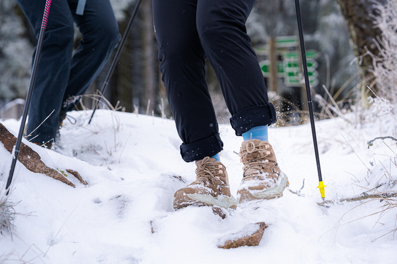 Schritt für Schritt im Schnee – Jeder Tritt zählt! Nahaufnahme von Wanderstiefeln, die durch tiefen Schnee stapfen. Die Füße gehören zu Wanderern mit schwarzen Hosen und blauen Socken. Wanderstöcke mit gelben Spitzen stecken im Schnee, während der Boden von Schnee und Felsen bedeckt ist.