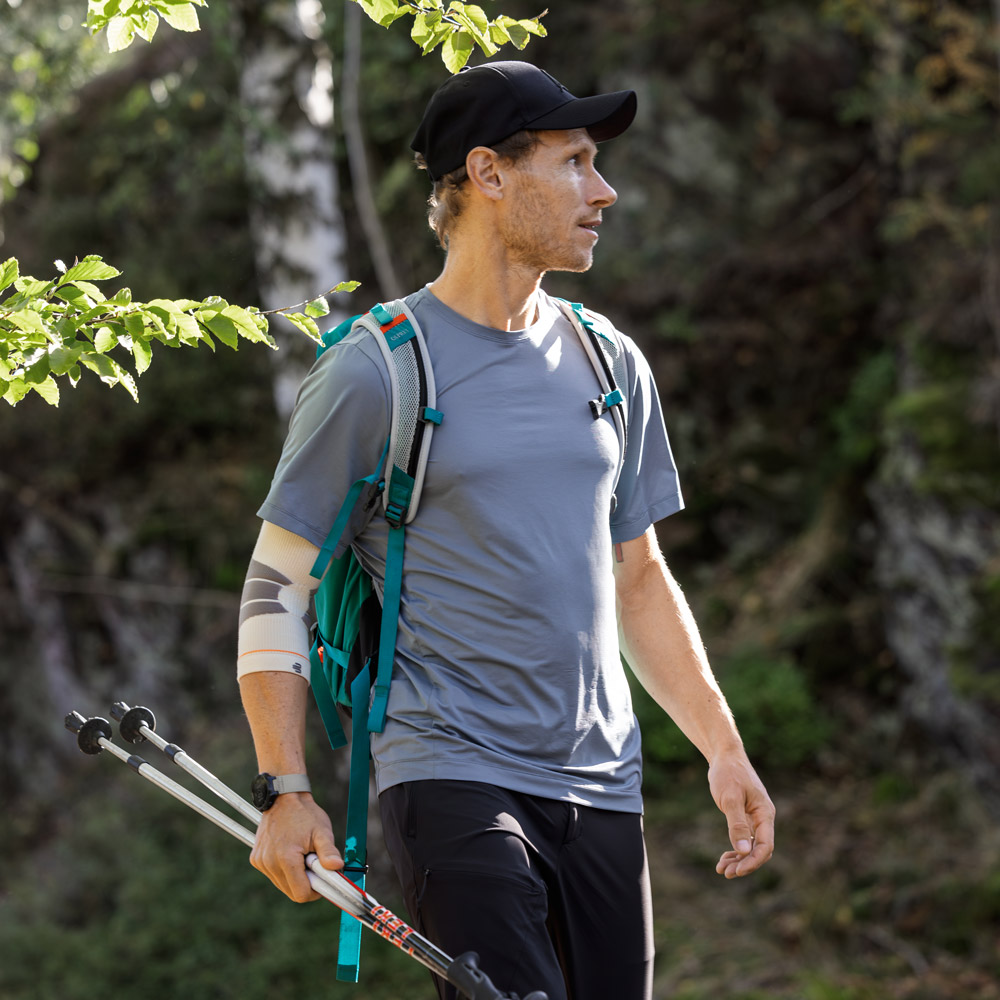 in Wanderer steht auf einem schattigen Waldpfad und blickt aufmerksam in die Umgebung. Er trägt einen Rucksack, hält Trekkingstöcke in der Hand und nutzt am linken Arm einen Outdoor Compression Elbow Support.