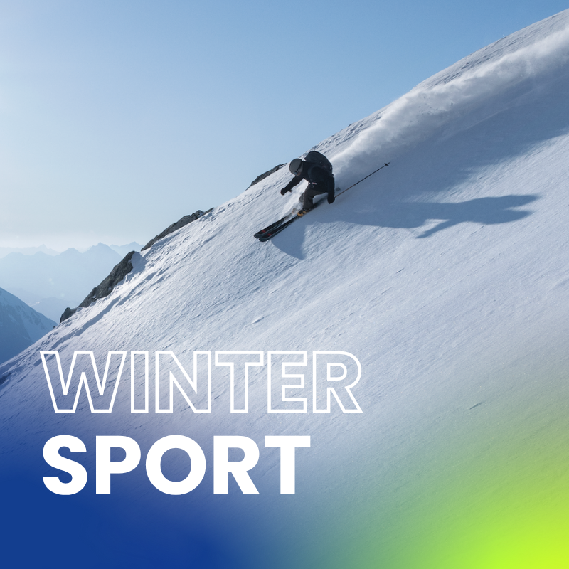 Skifahrer fährt in steiler Hanglage einen Tiefschneehang hinab, wirbelt Schnee auf und zeigt eine dynamische Abfahrt im alpinen Gelände bei klarem Himmel.