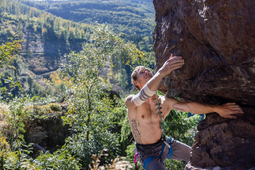 Weitwinkelaufnahme eines Kletterers an einem Felsüberhang mit Blick ins Tal; am Arm trägt er die Bauerfeind Sports Outdoor Compression Elbow Support in Sandstone, während er sich am Fels abstützt.