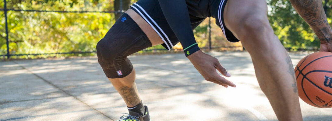 Basketballer mit schwarzer Kniebandage beim Dribbling Ein Basketballspieler trägt eine schwarze Kompressionsbandage am Knie und dribbelt den Ball auf einem Outdoor-Court. Die Bandage hebt sich deutlich vom restlichen Outfit ab und zeigt das NBA-Logo sowie das Logo des Herstellers.
