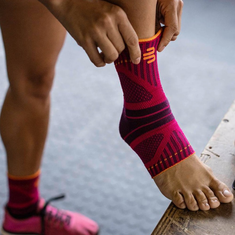 Schnell angezogen, optimal unterstützt! Nahaufnahme eines Fußes mit einer pink-schwarzen Sprunggelenkbandage. Eine Hand zieht die Bandage über das Sprunggelenk, während im Hintergrund ein rosa Sportschuh zu sehen ist.